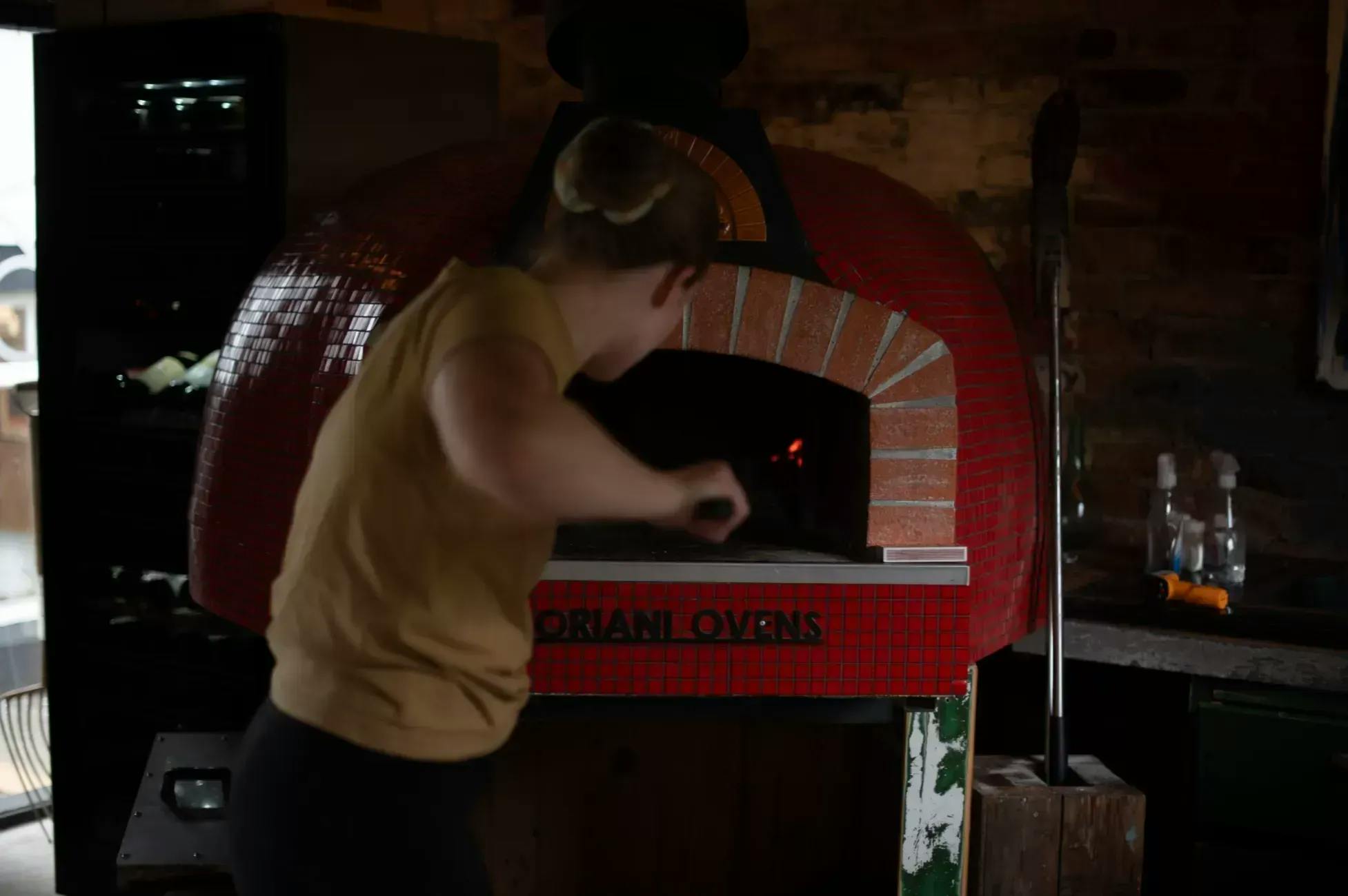 Jente som setter inn en pizza med pizzaspade i en rød italiensk pizzaovn. Merket på ovnen er Fiorni Ovens